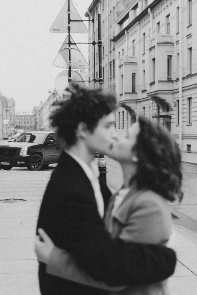 Una coppia si bacia nel centro città. I soggetti sono sfocati mentre è nitido lo sfondo cittadino. La foto è in bianco e nero.