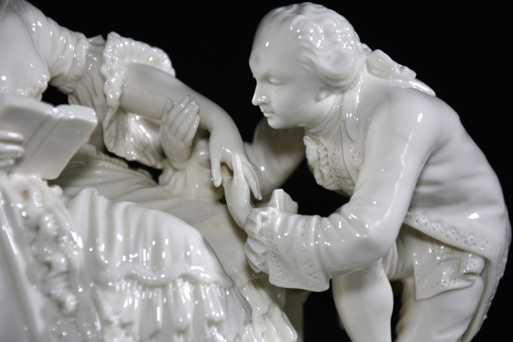 24000 baci -  Una statua di porcellana raffigurante una damina seduta con ampi abiti ottocenteschi, che porge la mano ad un gentiluomo con capelli lunghi ricci raccolti in un acoda, nell'intento di baciare la mano alla dama. la foto è in bianco e nero