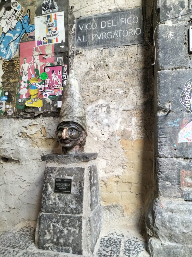 CITTÀ- centro storico. La foto ritrae la statua del volto di Pulcinella. Appoggiata su una pietra, l'opera realizzata in bronzo presenta un cappello a punta molto lungo, una maschera sugli occhi e un naso aquilino pronunciato. Quest'ultimo cattura l'attenzione perché è dorato e non color bronzo come il resto del volto.