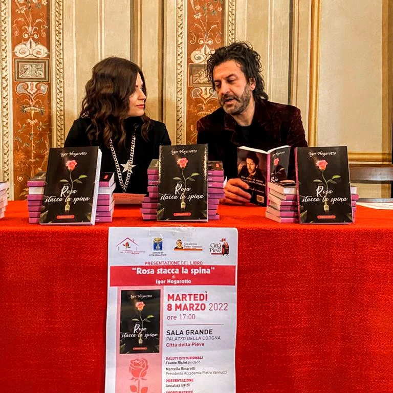 Rosa tour nazionale - Annalisa Baldi e Igor Nogarotto sono seduti al tavolo di conferenza in occasione della presentazione del libro. Annalisa ha lunghi capelli castani, indossa una maglia nera, sta guardando Igor parlare. Igor indossa giacca e camicia scura. Sul tavolo ci sono diverse copie del libro e il tavolo è coperto da una tovaglia lunga rossa. 