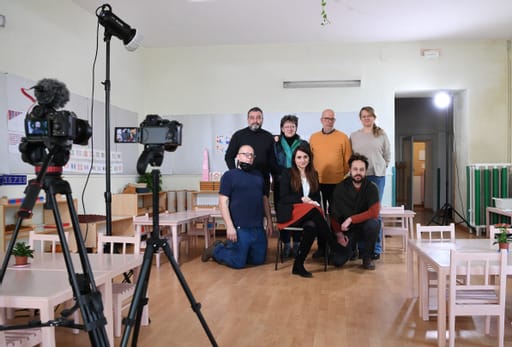 Gruppo di uomini e donne che curano il progetto Montessori Road. Si trovano all'interno di un'aula di una scuola Montessori, con arredi bianchi e pavimento in legno. Davanti a loro ci sono due telecamere e il microfono.