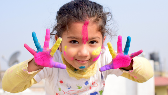 Una bambina con le mani colorate e la maglietta bianca sporca di colori sorride. Ha strice fucsia, gialle e blu in faccia e spalanca le manine vicine al viso.
