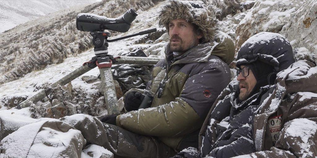 VINCENT MUNIER e SYLVAIN TESSON. Il fotografo e lo scrittore si trovano appostati tra le rocce innevate. Entrambe indossano giacche, cappello e guanti per proteggersi dal freddo. Vincent ha davanti a sé la telecamera e il tre piedi per scattare foto agli animali e ai paesaggi. 