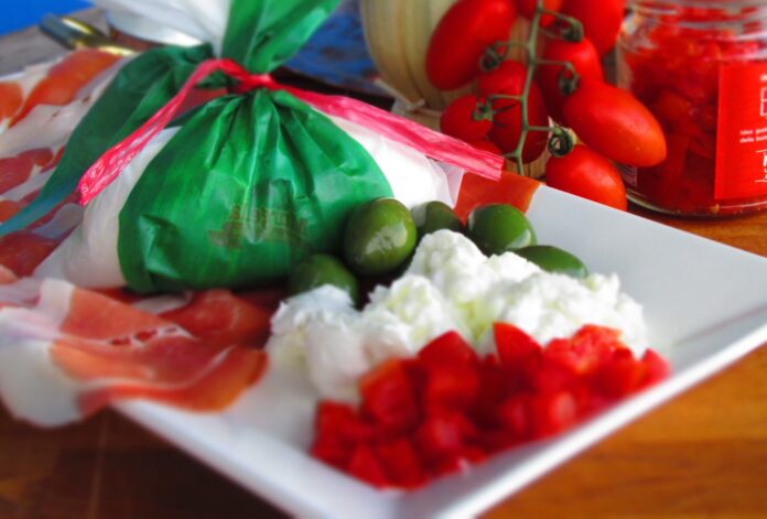 una burrata confezionata con carta tricolore italiana, posta su un piatto dove c'è anche della mozzarella e del pomodoro ttagliati a cubetti. sullo sfondo dei pomodorini e del cibo fresco