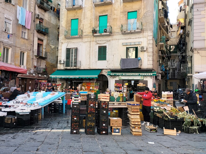 CITTÀ- Quartieri Spagnoli: Mercato della Pignasecca. Si tratta del mercato principale dei quartieri spagnoli e si trova in una piccola piazza circondata da condomini e negozi. L'immagine mostra a sinistra un banco del pesce mentre al centro e a destra ci sono cassette di plastica nera e di legno con all'interno frutta e verdura. Dietro le bancarelle ci sono tre condomini separati tra loro da due strette vie.