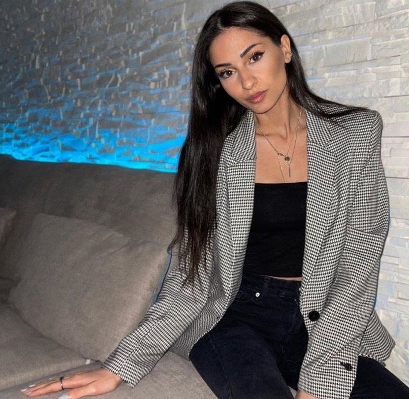 ella myeers indossa pantaloni e top nero scollato, sopra una giacca grigia. E' seduta su un divano di stoffa chiara