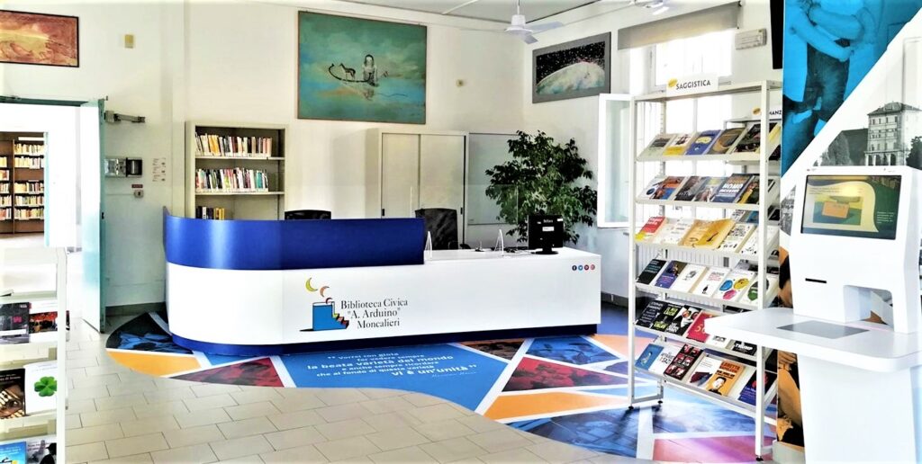 la biblioteca civica Arduino con in vista degli scaffali pieni di libri e il bancone della reception bianco con un vetro blu e illogo sul bancone