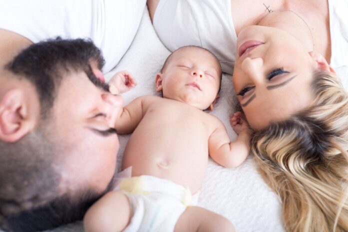 doppio cognome - nella foto un neonato con solo il pannolino dorme sul lettone in mezzo a mamma e papà che sono posizionati al contrario rispetto il bambino e lo guardano amorevolmente