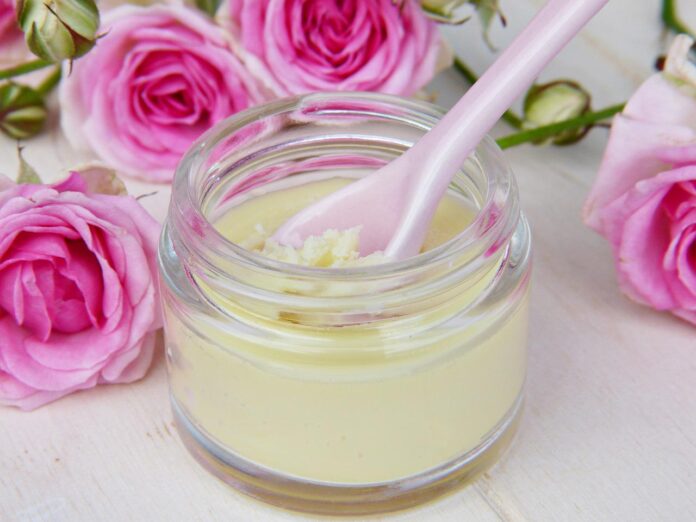 Burro di karitè, cosmetico naturale, pelle, capelli, utilizzo. Un barattolo in vetro con all'interno il burro di karitè. Dentro un cucchiaino di colore rosa e sullo sfondo delle rose.