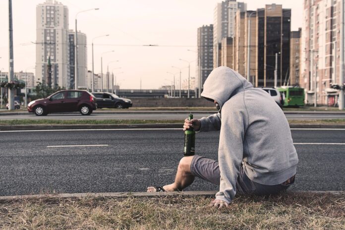 andrea strange - un uomo, che indossa pantaloni corti e una felpa grigia col cappuccio sulla testa, seduto sul ciglio della sstrada, con una bottiglia di birra in mano