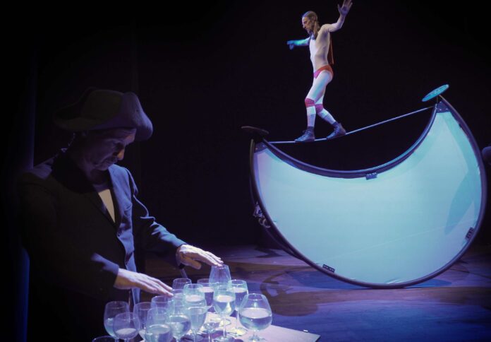 Torino Fringe FEstival - un artista cammina su una fune posizionata sulle due punte di un amezzaluna gigante bianca, mentre un altro artista con un cappello vistoso blu sfiora conle dita i bordi di alcuni bicchieri per farli suonare