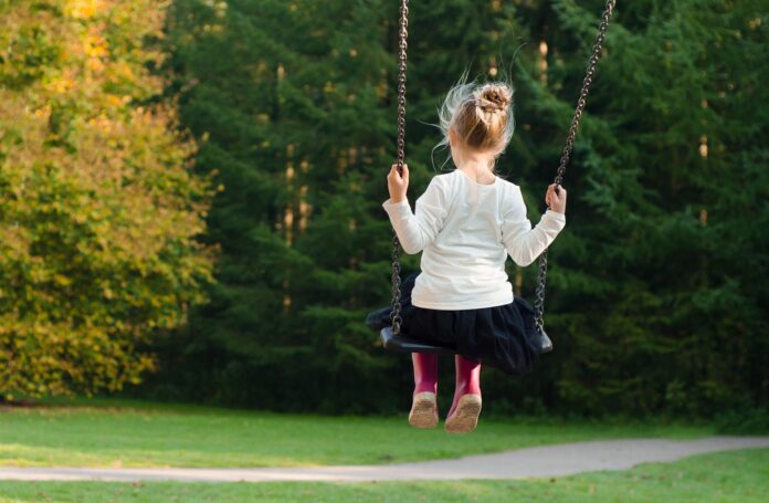 Una bambina con maglietta bianca, gonna nera, capelli biondi raccolti in una coda, è seduta su un'altalena nel parco
