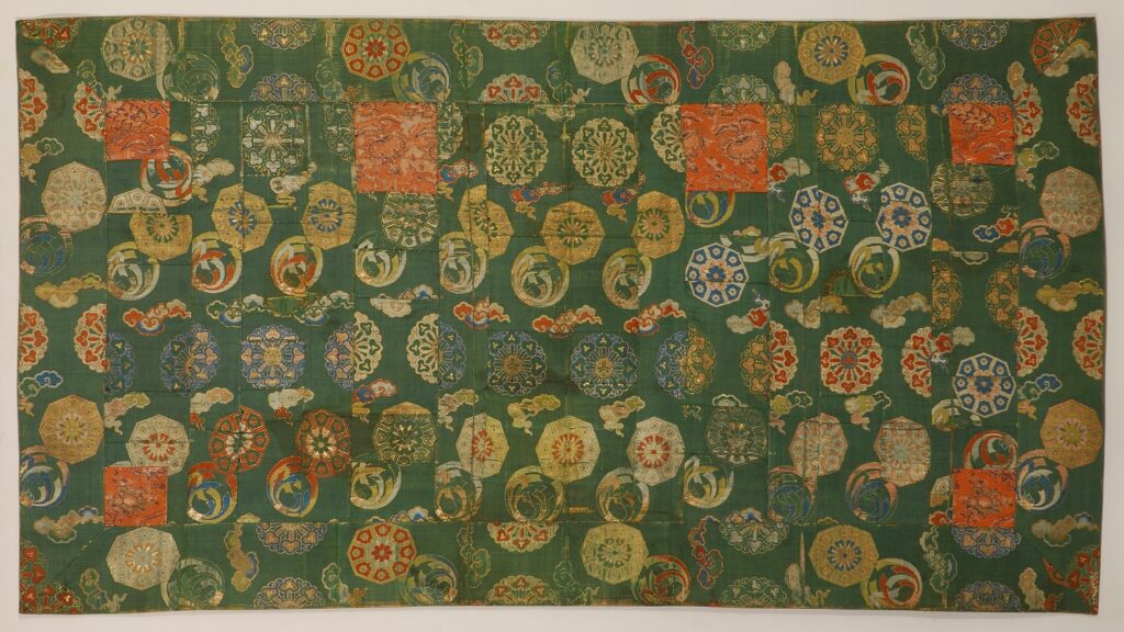 KESA- immagine del tessuto del primo kesa esposto al MAO. Il tessuto in seta verde presenta decorazioni minuziose color ocra, azzurro e rosso.