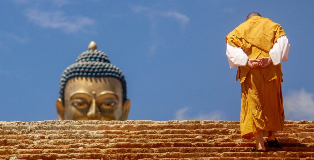 KESA- un monaco buddhista con indosso il kesa color ocra sale strette scale con le mani incrociate dietro la schiena. Da dietro le scalinate si vede la testa di una statua dorata del Buddha, che sembra osservare il monaco sulla destra dell'immagine. 