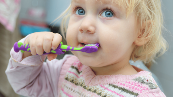 sigilaltura dei solchi dei denti molari in foto una bambina di pochi anni bionda con occhi azzurri uno spazzolino viola e verde in mano un maglioncino rosa bianco e verde