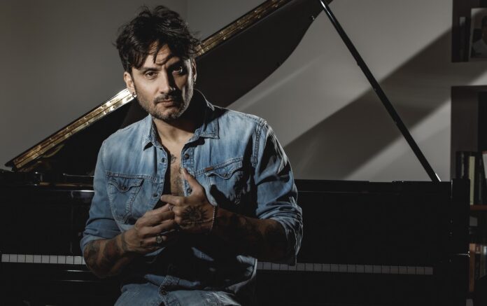 Fabrizio Moro seduto e dietro di lui un pianoforte a coda. Fabrizio veste una camicia di jeans aperta sul petto e si tocca gli anelli