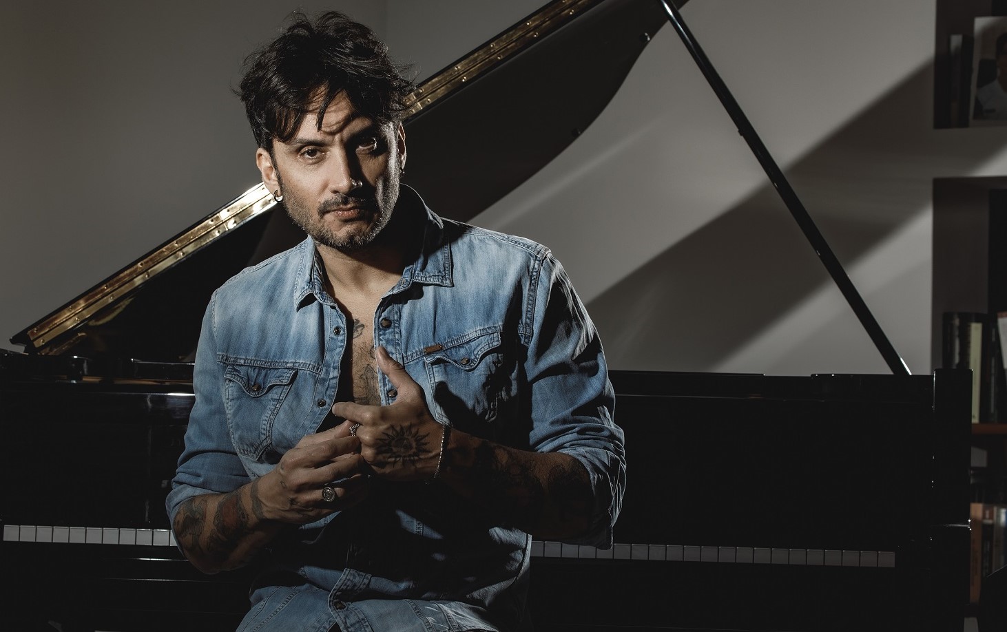 FABRIZIO MORO (© F🇮brizi🇮 Cest🇮ri)(2) Fabrizio Moro seduto e dietro di lui un pianoforte a coda. Fabrizio veste una camicia di jeans aperta sul petto e si tocca gli anelli