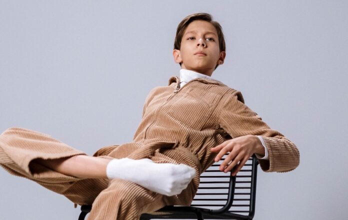 Parental abuse - un bambino vestito con un completo di pantaloni e giacca beige a righine, cmicia bianca, calini bianchi e senza scarpe, sieede con fare superbo su una sedia