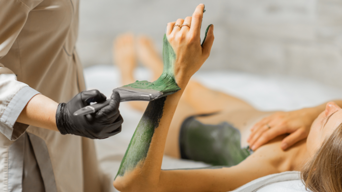 Fangoterapia, anticellulite, estetico, curativo. Una donna sdraiata sul lettino mentre le viene spalmato su un braccio del fango termale di colore verde intenso.