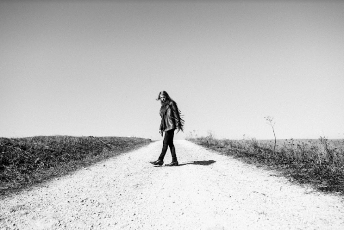 vision - black snake moan, jeans neri, stivali a punta e giacca di pelle scura con le frange, è ritratto nel mezzo di una stada sterrata (foto in bianco e nero).