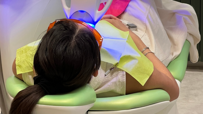 una donna si sottopone a un trattamento di odontoiatria estetica sdraiata su sedia dentista verde con occhiali da sbiancamento