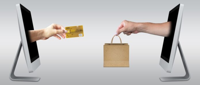 e-commerce - due schermi piatti di pc uno di fronte all'altro. Da uno sbuca una mano con una carta di credito tra le dita, dall'altro una ano con una busta di carta