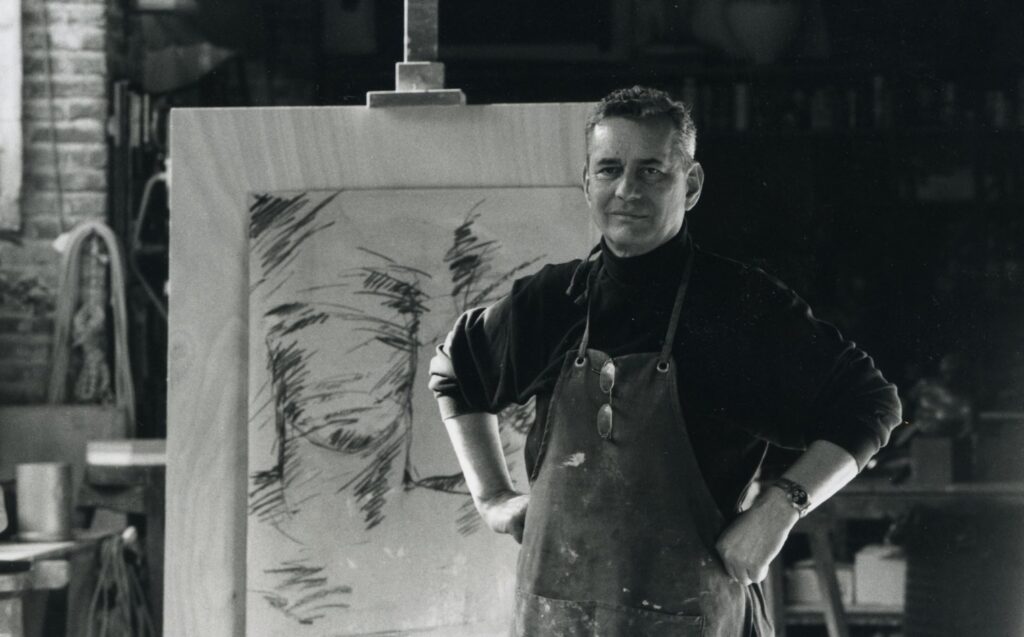 Mitoraj in una foto in bianco e nero, indossa un grembiule nero da pittore e al collo ha appeso degli occhiali dietro di lui un quadro con il disegno abbozzato di un volto
