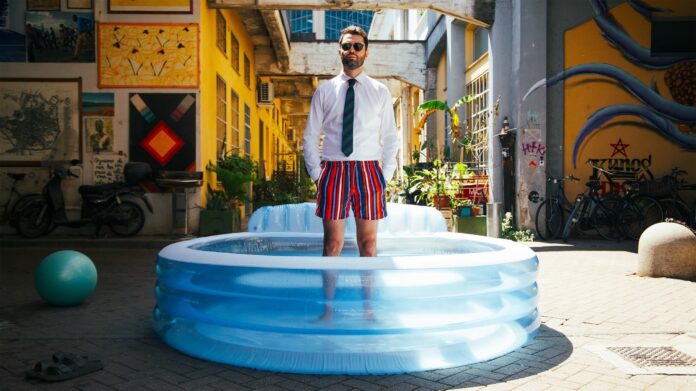 santoianni, che indossa costume a da bagno a riche, camicia bianca e cravatta, in piedi dentro a una piscina gonfiabile per bambini