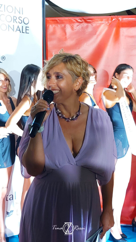 Miss Italia Liguria - la presentatrice della serata Stefania Vivioli con capelli corti biondi e mossi, indossa un abito color lavanda molto scollato, tiene in mano un microfono e sorride