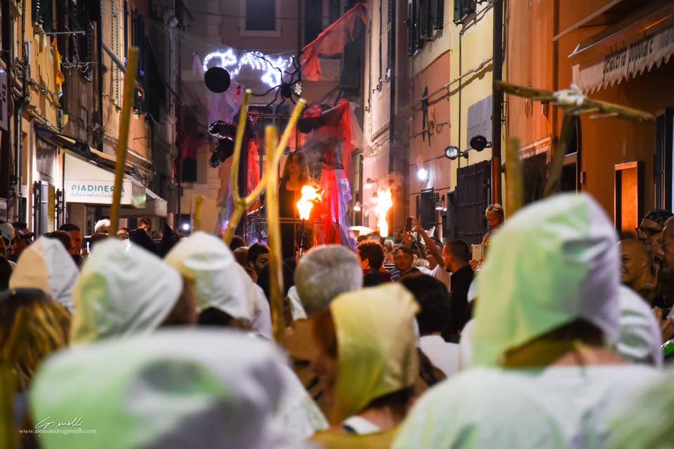 stregheFesta delle Basüe - nella foto tanta gente di schiena in un corteo. Sono vestiti dcon un mantello bianco con cappuccio e seguono un carro