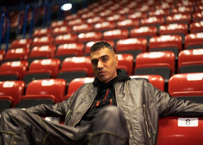 Marracash seduto da solo in un teatro di cui si vedono le poltroncine rosse disposte in salita