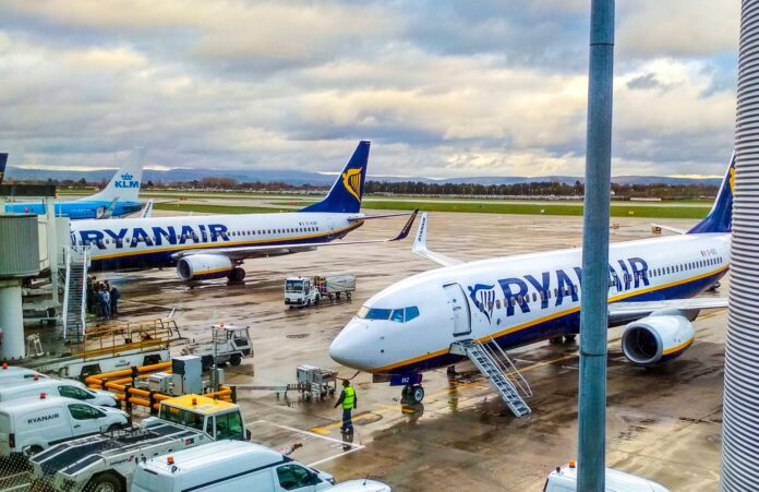 Ryanair voli low cost - nela foto due aerei bianchi con la scritta blu sono parcheggiati in un aeroporto. Intorno a loro degli operatori con giubbotto catarinfrangente e dei mezzi di lavoro