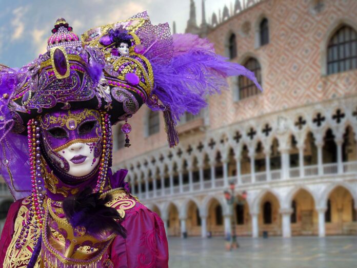 Venezia - sulla destra il palazzo ducale e in primo piano sulla sinistra, una persona indossa una sontuosa maschera di carnevale che copre totalmente il viso. La maschera è bianca e viola e comprende un copricapo con delle piume viola