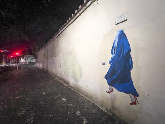 Kabul un anno dopo - su un muro di Roma il disegno di una donna afghana vestita con un abito blu e il burqa. La donna indossa scarpette rosse, simbolo della lotta contro la violenza sulle donne