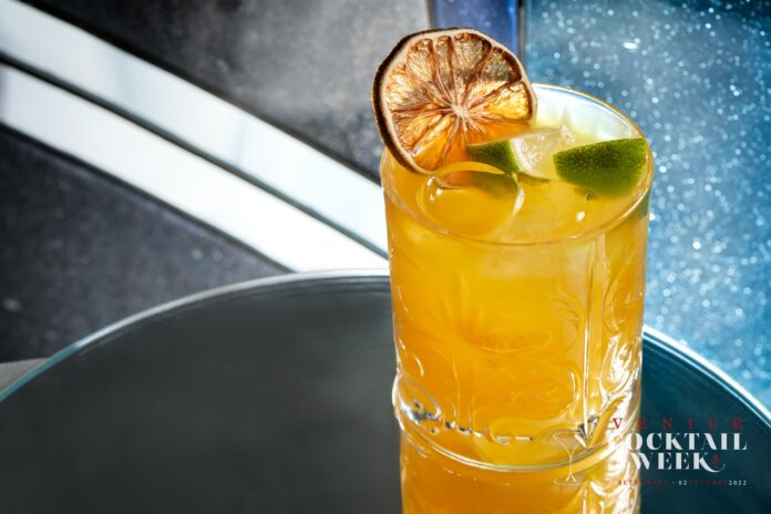 Venice Cocktail Week - un Bicchiere con un drink giallo, una fetta di limone secca e il logo della VCW
