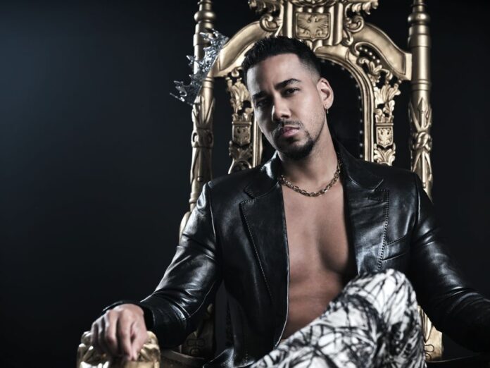 romeo santos in primo piano, seduto su un trono dorato, indossa pantaloni bianchi e neri, giacca di pelle. E' a torso nudo con una spessa collana d'oro al collo