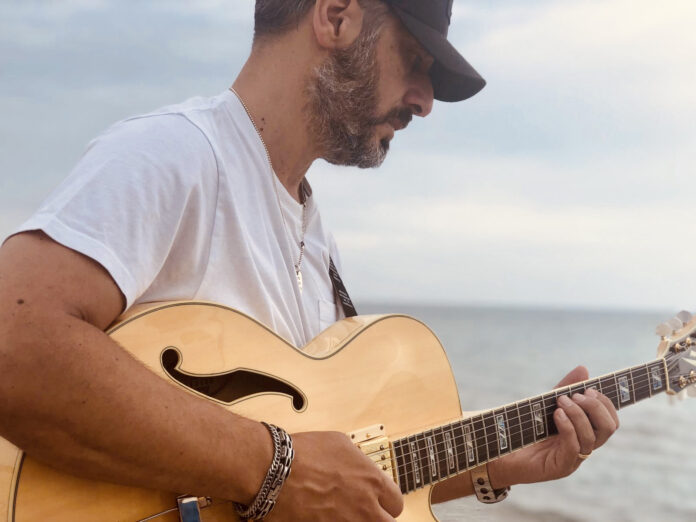 ambrosino in primo piano, indossa una tshirt bianca, cappellino scuro e imbraccia una chitarra acustica di legno chiaro