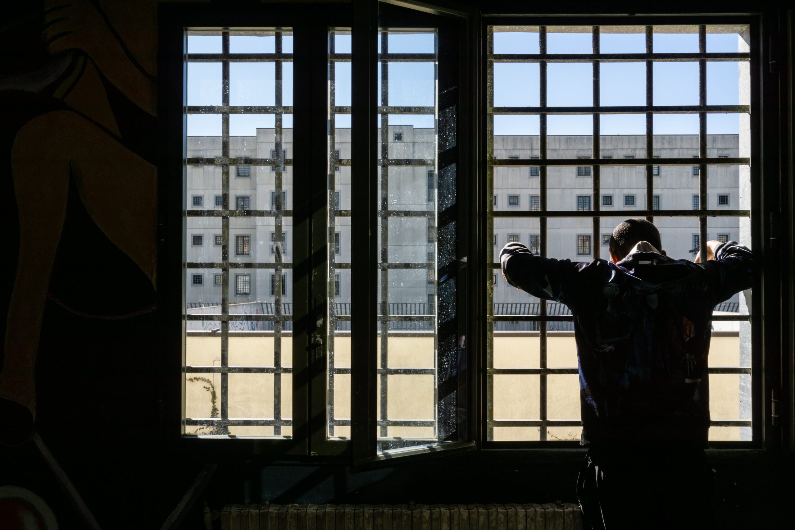 Ri-scatti - un ragazzo di spalle giarda un cortile da una vetrata protetta da sbarre