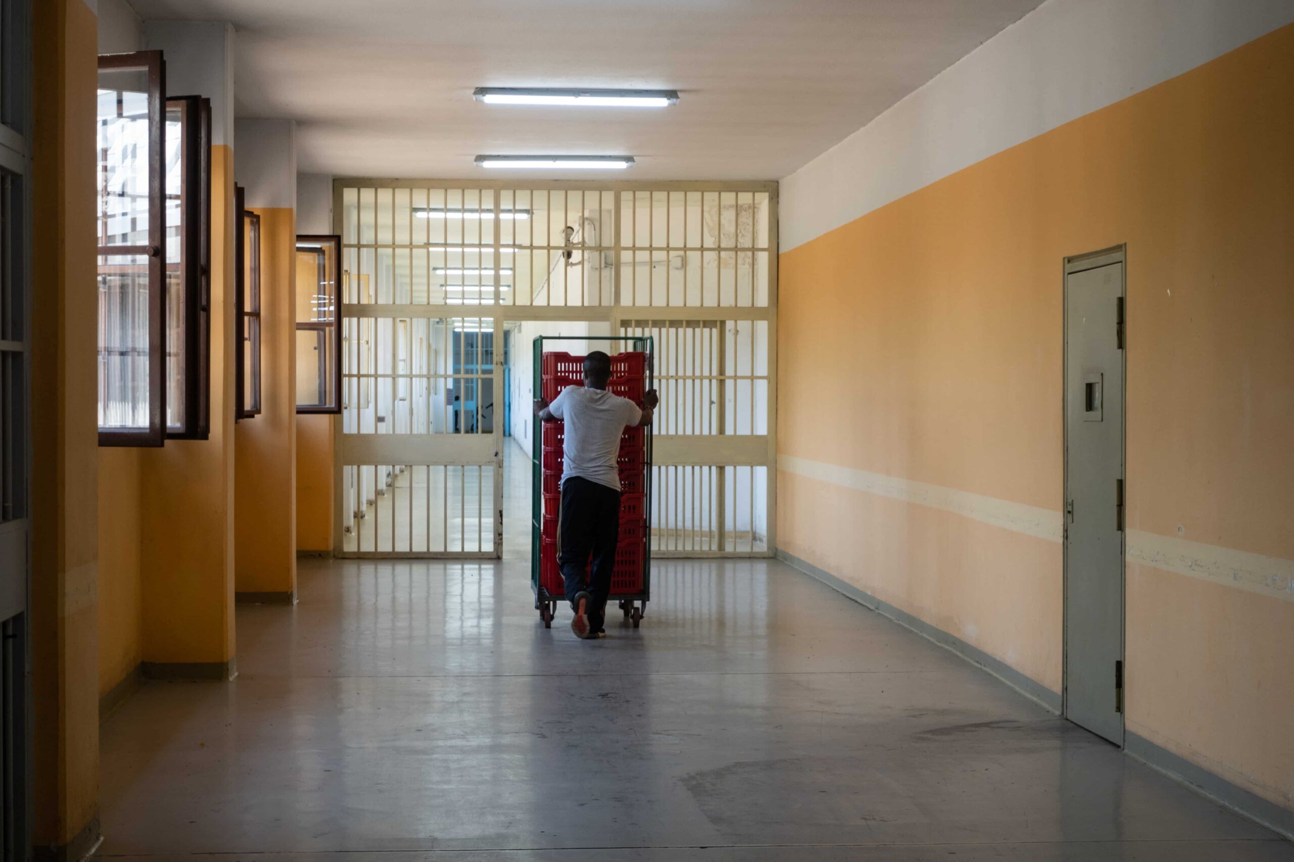 Ri-scatti - nella foto un ragazzo di schiena spinge una gabbia piena di cassette di plastica rosse, lungoun corridoio che al fondo ha delle porte a sbarre