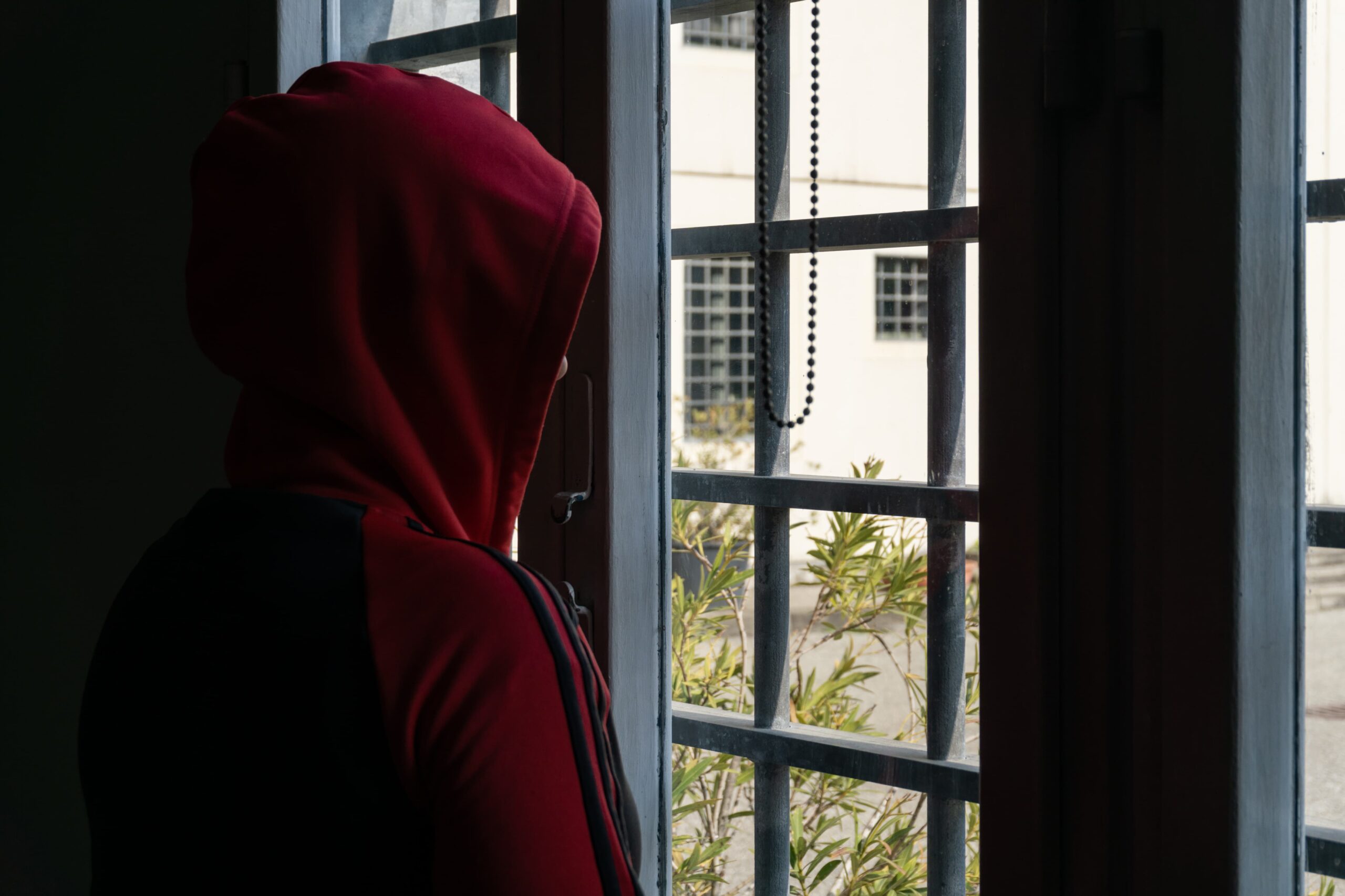 carcere - un ragazzo indossa una felpa con cappuccio rossa e guarda fun giardino fuori da una finestra con le sbarre