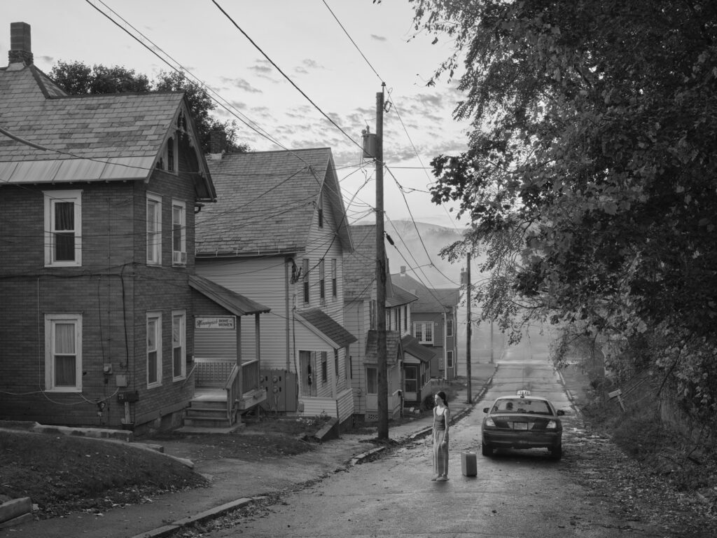 una donna i  piedi in una strada solitaria con auto sulla destra e case sulla sinistra bianco e nero