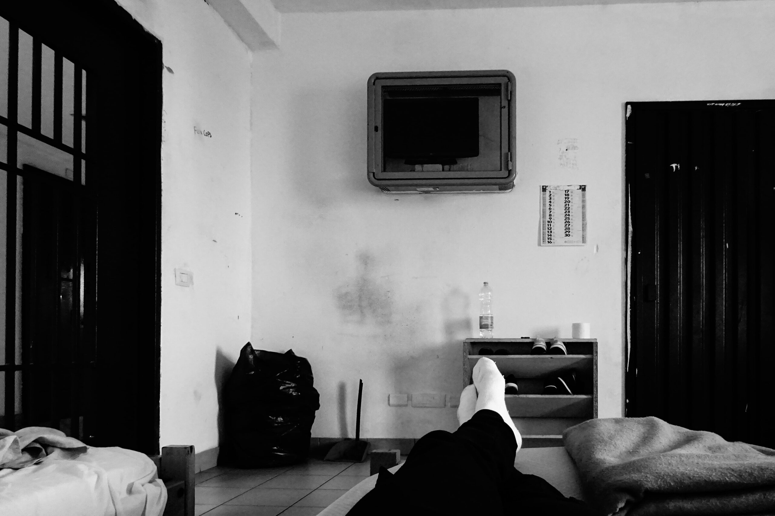 Carcere - foto in bianco e nero di un acella con dei mobili poveri, una branda su cui è diseso un deteneto di cui si vedono i piedi coperti da calzini bianchi