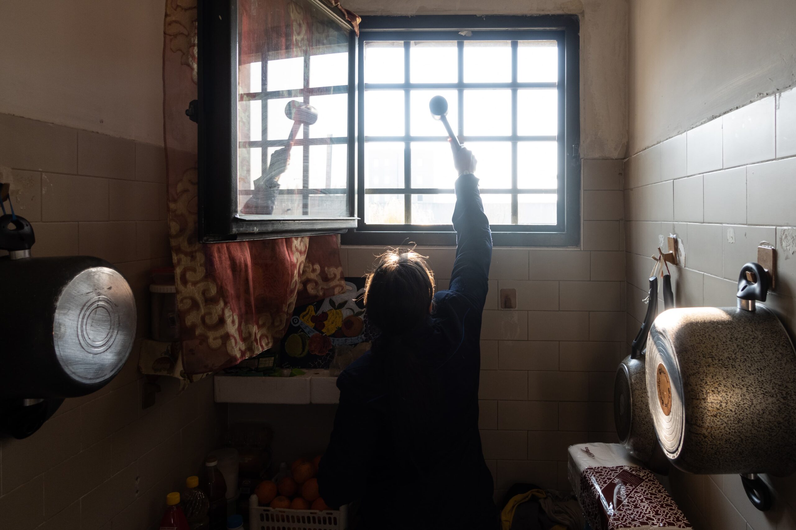 una donna dietro una finestra con le sbarre ha le braccia alzate e tiene in mano un mestolo