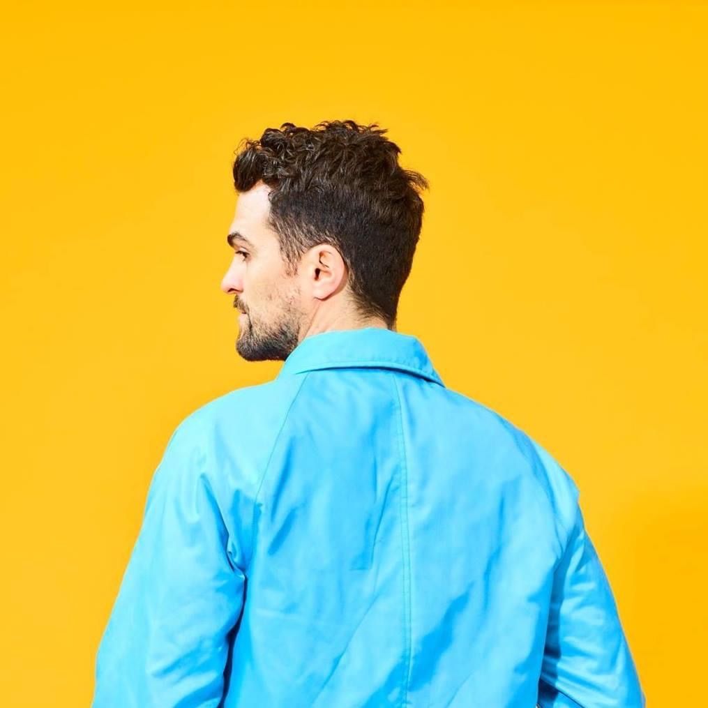 Scombinanto - street artist di spalle, con capelli e barbetta scura, indossa una camicia azzurra e lo sfondo è giallo