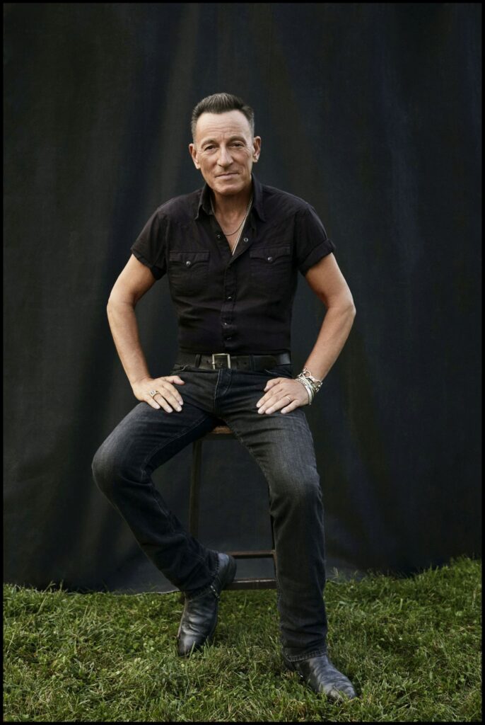 bruce springsteen seduto su uno sgabello, indossa camicia e jeans neri