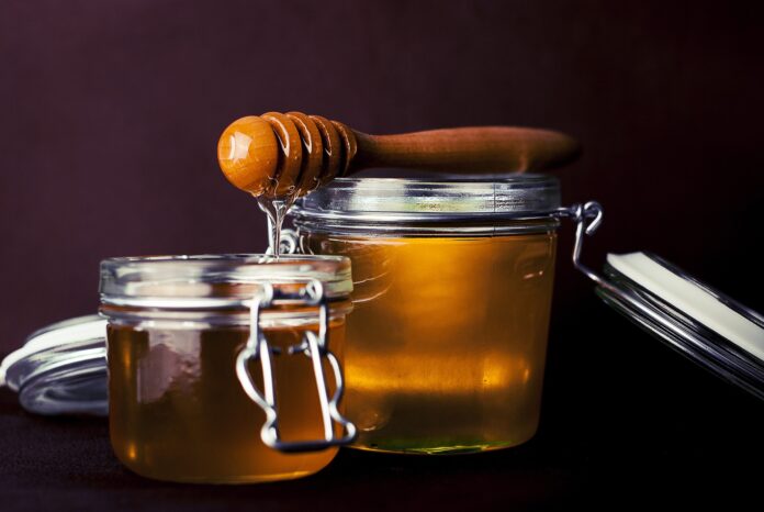 miele dentro a due vasetti di vetro e un martelletto da mieleappoggiatosopra un vasetto da cui cola il miele