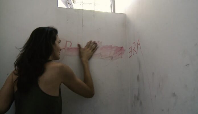 una donna cncella una scritta rossa progioneira dentro una stanza bianca