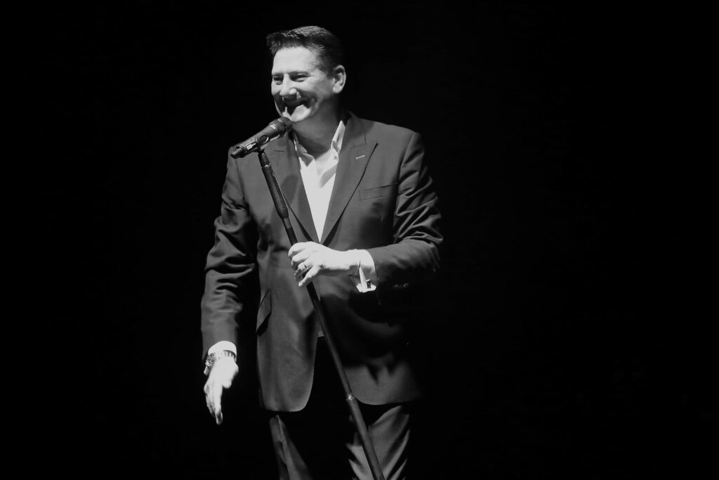 tony hadley in primo piano, indossa vestito scuro e camicia bianca, ha l'asta del microfono nella mano sinistra