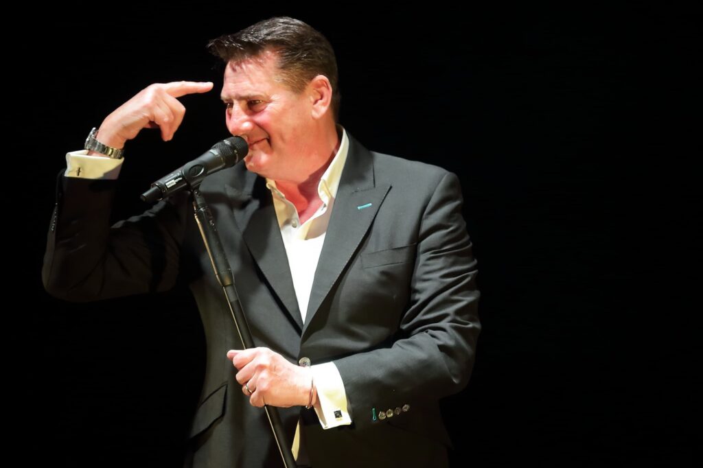 40th anniversary tour: Tony Hadley con giacca scura camicia bianca davanti al microfono ride e si porta una mano alla testa facendo il segno della pistola