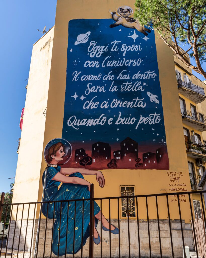 A Roma una facciata di un palazzo con un'opera di street art con scritta una poesia e sotto la poesia una donna seduta con un vestito azzurro lungo e la testa dentro un casco trasparente come quello degli astronauti